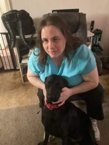 Angela, with long brown hair, dressed in a light blue shirt and black pants, sits on a chair and gently cradles a black English Labrador Service Dog named Steve with a red collar.